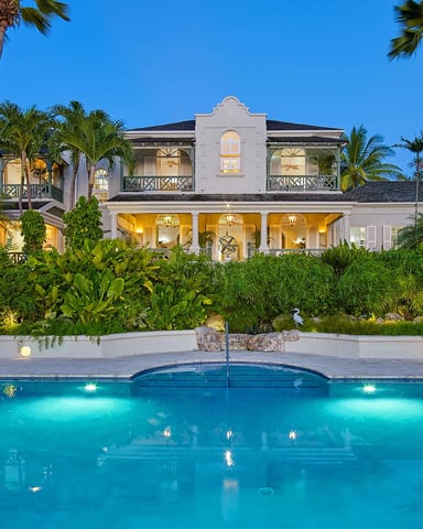 Two-story white colonial house with illuminated windows, surrounded by palm trees and tropical landscaping, viewed from lit swimming pool at dusk