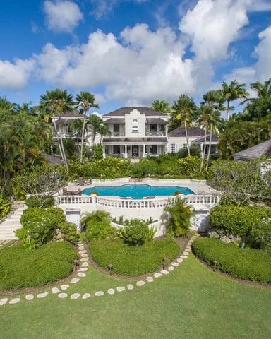 White two-story colonial house with pool, manicured lawn, palm trees, and stone pathways in tropical setting.
