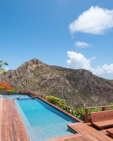 Hillside villa with infinity pool, wooden deck, lounge chairs, and panoramic mountain views under clear blue sky