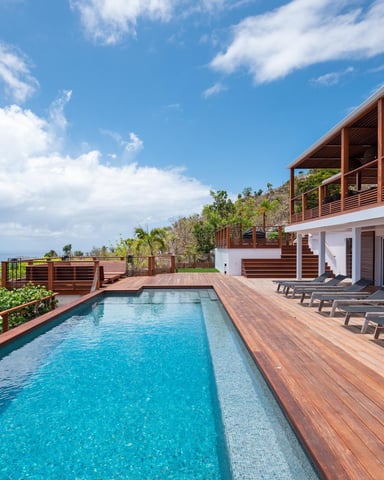 Modern hillside villa with infinity pool, wooden deck, lounge chairs, and ocean view under blue sky