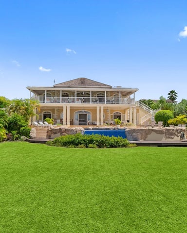 Two-story colonial-style house with wraparound porch, pool, and manicured lawn surrounded by tropical landscaping