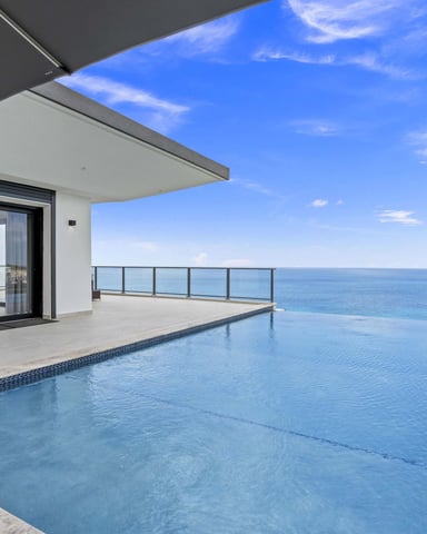 Modern oceanfront terrace with infinity pool, black umbrella, glass railing, and clear blue sea view
