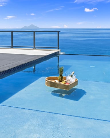 Infinity pool overlooking blue ocean with distant volcanic island, floating basket with pineapple and drinks, egg chair on deck