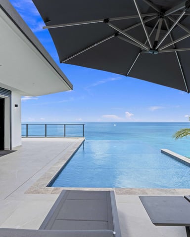 Oceanfront terrace with infinity pool, black umbrella, lounge chairs, and sea view beyond glass railing