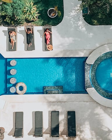 Aerial view of resort pool and hot tub with three guests sunbathing on lounge chairs, surrounded by palm trees and manicured lawn.