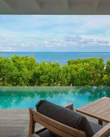 Covered deck with lounge chairs overlooking infinity pool, tropical vegetation, and turquoise ocean beyond