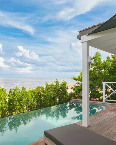 Beachfront deck with infinity pool, wooden lounge chairs, white pergola, and ocean view beyond mangrove vegetation