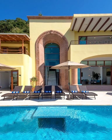 Modern yellow villa with swimming pool, lounge chairs with umbrellas, and arched entrance overlooking hillside