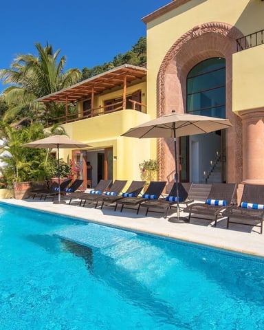 Yellow villa with swimming pool, lounge chairs, umbrellas, and palm trees under clear blue sky