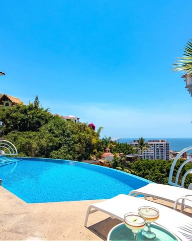 Modern white villa with blue swimming pool, lounge chairs, tropical plants, and ocean view under clear sky