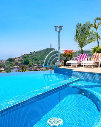Rooftop pool with blue water, striped loungers, palm trees, and coastal city view under clear sky