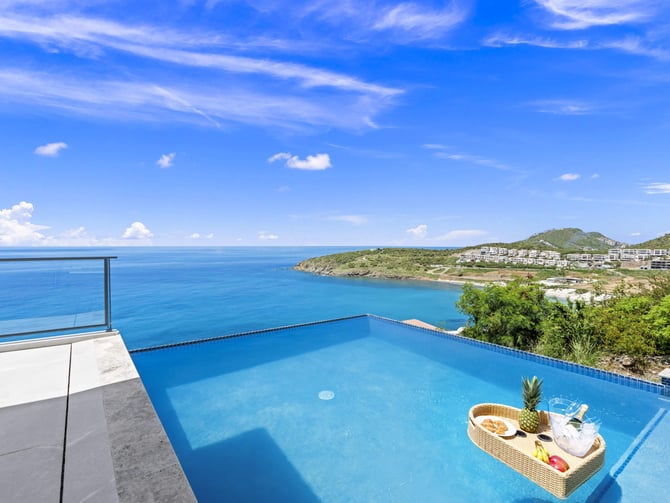 Infinity pool overlooking turquoise bay with coastal hillside development and white clouds in blue sky